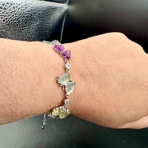 May include: A silver bracelet featuring a series of gemstones in shades of purple, green, and clear. The bracelet is composed of multiple gemstone clusters and is worn on a wrist.