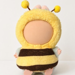 May include: A bee-shaped plush toy. It features a yellow hooded outfit with brown stripes, small pink hands and feet, and two yellow antennae. The face is a light peach colour.