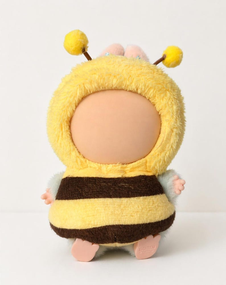 May include: A bee-shaped plush toy. It features a yellow hooded outfit with brown stripes, small pink hands and feet, and two yellow antennae. The face is a light peach colour.