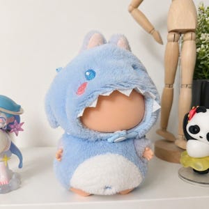 May include: A soft toy doll dressed in a light blue shark costume, featuring a white belly and a bow tie. The shark outfit has a white mouth and pink and blue details. The doll is displayed on a white shelf with other figures.