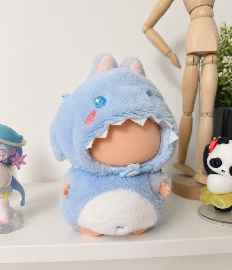 May include: A soft toy doll dressed in a light blue shark costume, featuring a white belly and a bow tie. The shark outfit has a white mouth and pink and blue details. The doll is displayed on a white shelf with other figures.