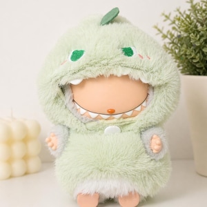 May include: A soft toy doll wearing a light green dinosaur costume with a hood, embroidered eyes, and a small leaf. The doll has a white-toothed grin and small hands and feet. The costume is fluffy and soft.