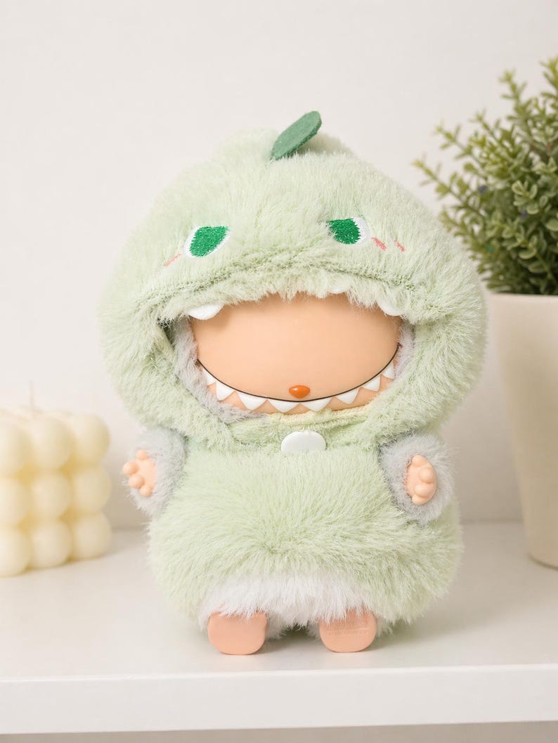 May include: A soft toy doll wearing a light green dinosaur costume with a hood, embroidered eyes, and a small leaf. The doll has a white-toothed grin and small hands and feet. The costume is fluffy and soft.