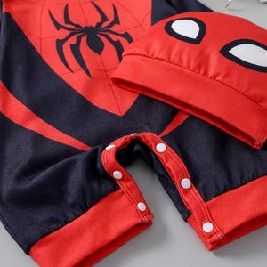 Op de afbeelding: Een rood en zwart baby-outfit met bijpassende hoed. De outfit heeft een groot zwart spinnenembleem op de borst en een spinnenwebontwerp. De hoed heeft witte oogvormen. De outfit heeft witte drukknoopsluitingen.