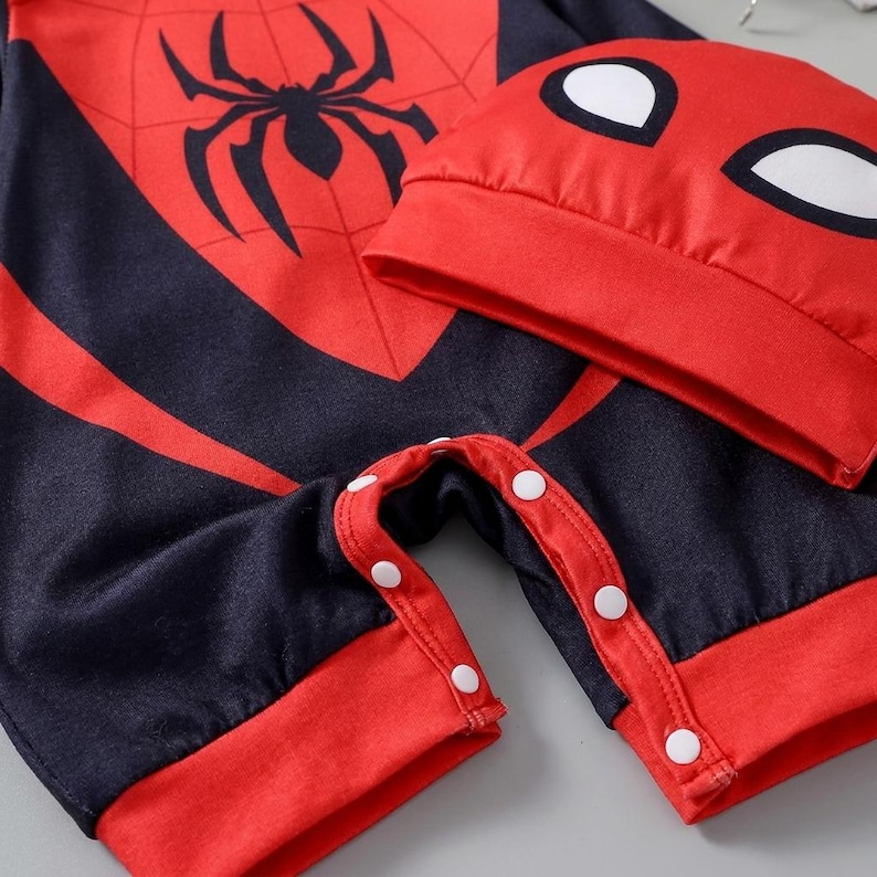 Op de afbeelding: Een rood en zwart baby-outfit met bijpassende hoed. De outfit heeft een groot zwart spinnenembleem op de borst en een spinnenwebontwerp. De hoed heeft witte oogvormen. De outfit heeft witte drukknoopsluitingen.