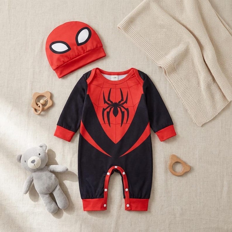 Op de afbeelding: Een rode en zwarte babyromper met een spinnenembleem, lange mouwen en drukknoopsluitingen. Een bijpassende rode hoed met witte oogvormen is inbegrepen. Een grijze teddybeer en houten speelgoed staan ook op de foto.