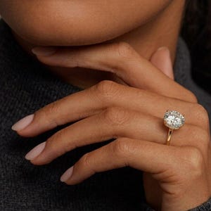 Può includere: Un anello in oro con una grande pietra preziosa trasparente a taglio brillante rotondo, montata su una struttura a quattro griffe. La fascia è sottile e in oro lucido, e la montatura include un delicato alone di pietre più piccole e luminose.