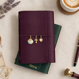 Peut inclure: Un carnet en cuir violet foncé avec les initiales KS gaufrées sur la couverture, orné de trois breloques dorées : un coquillage, une croix et un cœur. Il repose sur un carnet en cuir vert foncé avec le nom Olivia gaufré en lettres dorées.