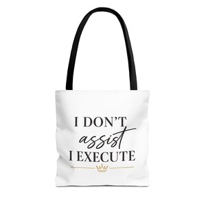 Könnte beinhalten: Weiße Tragetasche mit schwarzen Henkeln. Der Text "I DON'T assist I EXECUTE" ist in schwarzer Schrift geschrieben, darunter eine goldene Krone. Zum Tragen von Gegenständen.