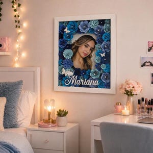 May include: A framed portrait featuring a young woman surrounded by blue paper flowers and butterflies. The name "Mariana" is written in cursive at the bottom. The frame is white, and the overall aesthetic is decorative and feminine.
