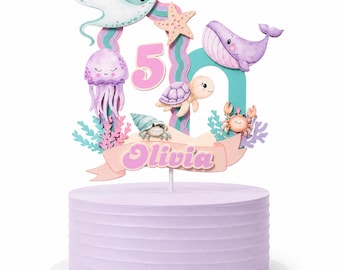 May include: A cake topper featuring a light purple number 5 and a banner with the name Olivia in pink letters. The design includes a stingray, jellyfish, whale, sea turtle, crab, and starfish in pastel colors, all set on a light purple cake.