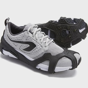 May include: A light gray athletic sneaker featuring a black rubber traction attachment over the sole and toe area. The shoe has a mesh upper, white midsole, and black laces. The traction device includes small metal studs for grip on slippery surfaces.