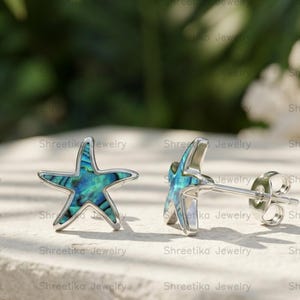 Puede incluir: Dos pendientes de botón con forma de estrella de mar. Los pendientes tienen un contorno plateado y una incrustación de concha de abulón azul-verde. Los pendientes se muestran sobre una superficie de piedra clara con un fondo verde borroso.