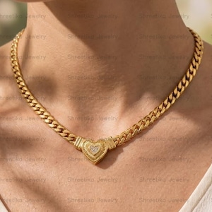 May include: A gold-toned curb chain necklace featuring a central heart-shaped pendant with a ribbed texture and a small cluster of clear stones set in the middle. The necklace rests against the neck, showcasing a polished metallic finish throughout.