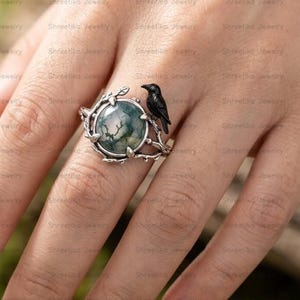May include: A silver ring featuring a round moss agate gemstone with green inclusions, set within a band designed as intertwined branches with small leaves and a small black bird perched on the side of the stone setting.
