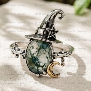 Op de afbeelding: Een zilverkleurige ring met een ovale mosagaat edelsteen als middelpunt. De zetting bevat een heksenhoedontwerp met een klein sterretje, een goudkleurig maansikkelaccent en takachtige details langs de zijkanten van de ringband.