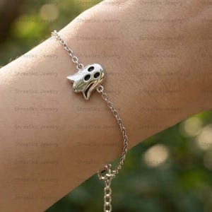 Op de afbeelding: Een zilverkleurige metalen kettingarmband met een centrale bedel in de vorm van een geest. De bedel heeft een glad, gepolijst oppervlak met twee donkere, ovale uitsparingen voor de ogen en een kleine, ronde opening voor de mond.