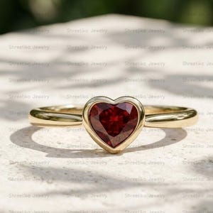 May include: A gold ring featuring a single heart-shaped dark red gemstone set in a smooth bezel setting. The band is polished yellow gold with a rounded profile. The gemstone is faceted and centered, creating a clear focal point on the ring.