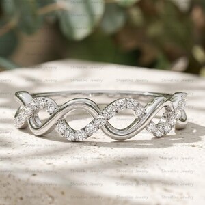 May include: A silver-toned ring featuring an infinity-style twisted band design. The band is accented with a series of small, clear, round-cut stones set along the intertwining metal loops, creating a continuous sparkling effect across the front.