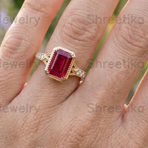 May include: A gold ring featuring a rectangular emerald-cut red gemstone set in a bezel with milgrain detailing. The band includes small white accent stones along the shoulders, creating a detailed design that highlights the central red stone.