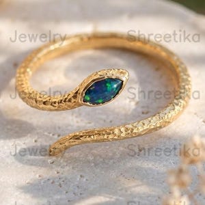 May include: A gold-toned ring shaped like a coiled snake with a textured, hammered surface. The snake's head features a marquise-cut dark blue opal gemstone with iridescent green flecks. The ring has an open, adjustable design.