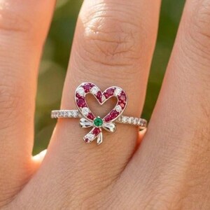 Op de afbeelding: Een zilverkleurige ring met een hartvormig zuurstokontwerp versierd met kleine rode en witte steentjes. Een enkele groene steen is gezet in het midden van een strik aan de basis van het hart, en de ringband is ingelegd met heldere steentjes.