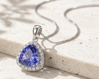 May include: A silver necklace featuring a trillion-cut violet-blue tanzanite gemstone pendant surrounded by a halo of small round white diamonds. The pendant is attached to a delicate silver box chain, showcasing a polished metallic finish.
