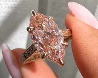 May include: A marquise-cut peach-colored gemstone ring set in a gold band. The band features intricate engraved floral detailing along the sides. The large, elongated stone is secured by six prongs, showcasing a faceted surface that reflects light.