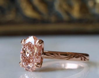 May include: A rose gold ring featuring a large, oval-cut peach-colored gemstone held by four prongs. The metal band is engraved with a delicate, continuous leaf pattern that wraps around the shank, showcasing a polished finish throughout.