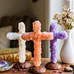 Könnte beinhalten: Drei stehende Kreuzskulpturen aus texturiertem, kristallinem Material in Weiß, Orange und Violett. Jedes Kreuz ist auf einem runden, dunkelbraunen, texturierten Sockel montiert. Die Kreuze sind in einer Reihe auf einer Holzoberfläche angeordnet.