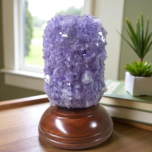 May include: A cylindrical lamp featuring a cluster of translucent purple amethyst crystals mounted on a smooth, circular, dark brown wooden base. The crystalline structure displays varied facets and jagged edges, creating a textured surface.
