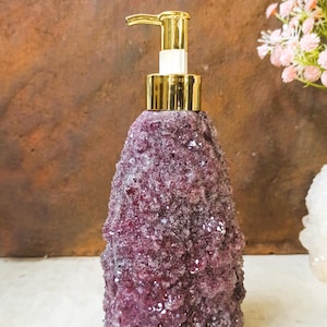 May include: A soap dispenser featuring a textured, deep purple amethyst crystal exterior and a polished gold-tone metal pump mechanism. The body of the dispenser has a rough, crystalline surface with natural variations in colour and light reflection.