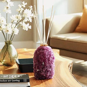May include: A purple crystal-like diffuser with white reeds, a gold ring, and a textured surface. The diffuser sits on a wooden table, next to a vase of white flowers and a black square container. Home decor.