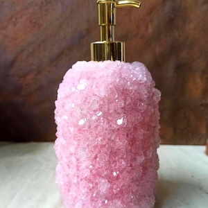 May include: Pink crystal-covered soap dispenser with a gold-colored pump. The dispenser has a cylindrical shape and is covered in sparkling, light pink crystals. The pump is a shiny gold color. The background is a blurred brown.