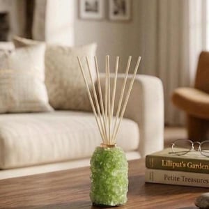 Puede incluir: Un difusor de varillas con una base cilíndrica hecha de material cristalino de color verde claro texturizado. Ocho varillas de madera delgadas, rectas y de color claro están insertadas en la parte superior de la base, abriéndose en abanico.