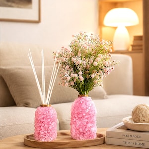 May include: Two cylindrical glass vases covered in small pink crystals. One vase holds several thin white reed diffuser sticks, while the other contains a bouquet of light pink baby's breath flowers. Both sit on a round wooden tray.