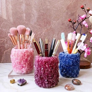 Puede incluir: Tres recipientes cilíndricos con una superficie cristalina texturizada en tonos rosa claro, magenta intenso y azul oscuro. Los recipientes contienen varios pinceles de maquillaje, bolígrafos y cepillos de dientes, junto a pequeñas piedras decorativas.