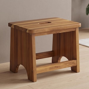 May include: A rectangular wooden stool crafted from medium-brown timber with a visible grain pattern. The seat features a centered oval cutout handle, supported by four sturdy legs connected by lower horizontal stretchers with arched cutouts.