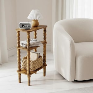 May include: A three-tiered wooden side table with turned legs, holding a white alarm clock, a small lamp, books, and a woven basket. The clock displays 12:59. A white armchair is to the right.