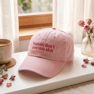 Könnte beinhalten: Hellrosa Baseballkappe mit dem gestickten Text "Women don't owe you shit" in dunklerem Rosa. Die Kappe liegt auf einer weißen Oberfläche, mit einer Kaffeetasse und einer Vase mit Rosen im Hintergrund.