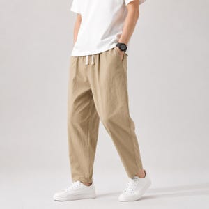 Könnte beinhalten: Beige Hose mit lockerer Passform, elastischem Bund mit weißem Kordelzug und Seitentaschen. Die Hose wird mit einem schlichten weißen T-Shirt, einer schwarzen Armbanduhr und weißen Turnschuhen kombiniert, was einen neutralen Look ergibt.
