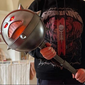 May include: A metallic gray mace with a round head featuring a menacing face with glowing red eyes and a wide grin. The mace has sharp spikes and a textured handle, suggesting a fantasy or cosplay item.
