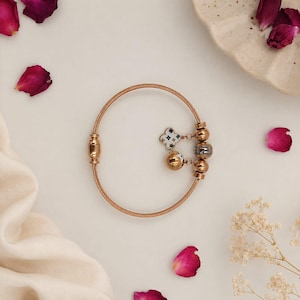 May include: A rose gold bracelet with a cable design. The bracelet features decorative charms, including a four-leaf clover and spherical beads with blue and white accents. The bracelet is displayed on a white surface with scattered rose petals.