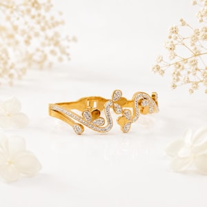 May include: A gold bracelet with a floral design, adorned with small, sparkling stones. The bracelet has an open design with curved lines and flower-like shapes. The background is white with soft, blurred floral accents.