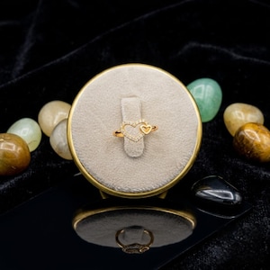 May include: A gold ring featuring a heart design. The main heart is encrusted with small, clear stones, with a smaller heart beside it. The ring is presented on a beige stand with a gold rim. The ring is a piece of jewellery.