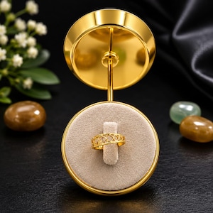 May include: A gold-colored ring with a heart-shaped design is displayed on a gold-colored ring stand. The ring is set on a beige, circular platform. The stand has a circular top and base, and a slender stem. The background is black.