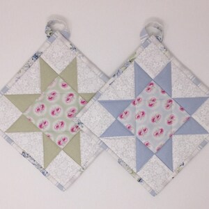 May include: Two quilted pot holders with a star design. One pot holder is green and white with a floral pattern in the center. The other pot holder is blue and white with a floral pattern in the center.