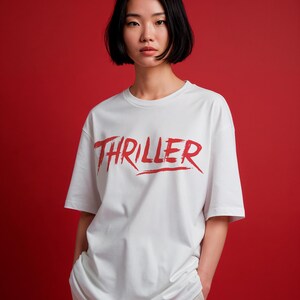 May include: White t-shirt with the word "THRILLER" in red, brush-style lettering. The shirt has short sleeves and a crew neck. The background is a solid red color.