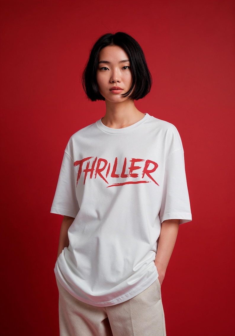 May include: White t-shirt with the word "THRILLER" in red, brush-style lettering. The shirt has short sleeves and a crew neck. The background is a solid red color.