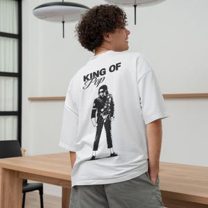 May include: White t-shirt with a black graphic of a person and the text "KING OF POP". The shirt is a short-sleeved, oversized style. The person in the image is wearing gray shorts.